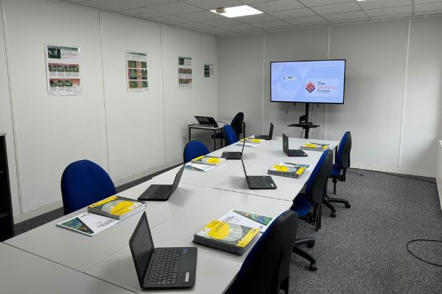 a training classroom set up with a long table with laptops and books all facing a screen at the front